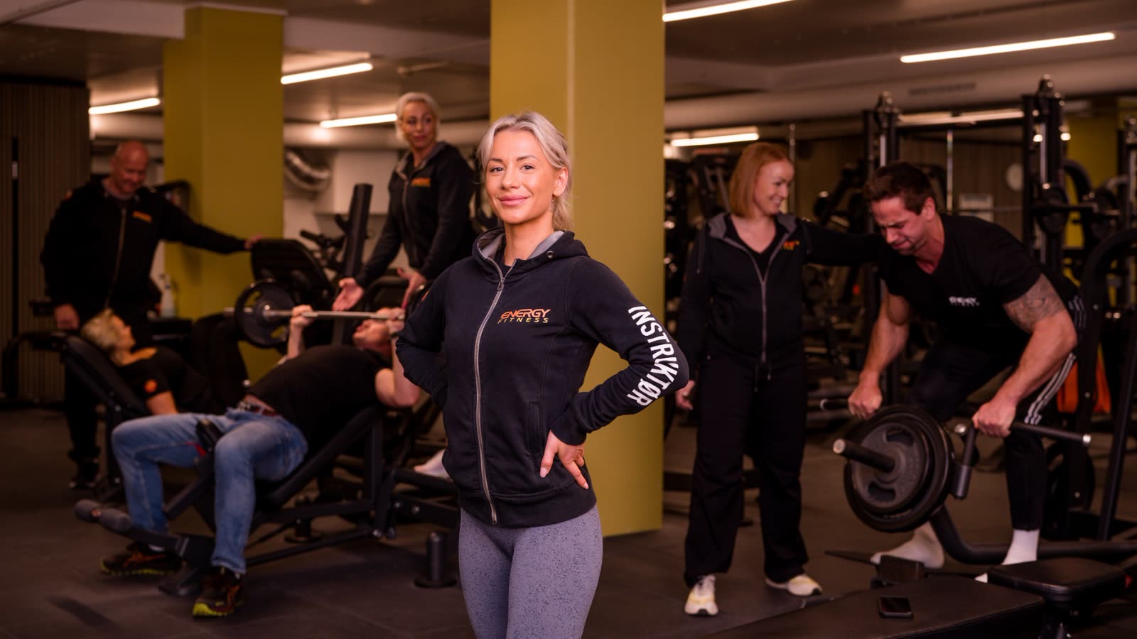 Instruktør i ENERGY Fitness-jakke foran medlemmer som trener på et treningsgulv